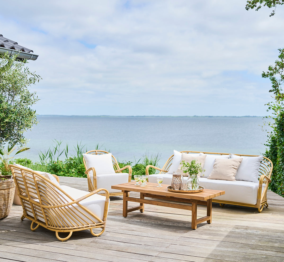 This is a lifestyle image of Julian Coffee Table in outdoor teak. On a seaside deck with lounge sofas and white cushions overlooking the ocean.