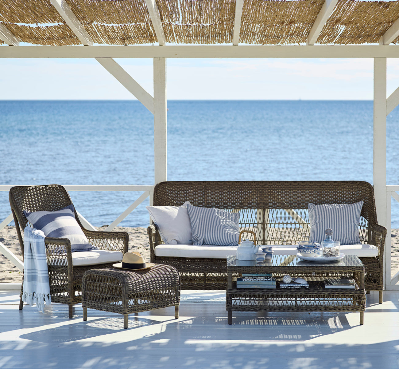 This is a lifestyle image of Hazel Coffee Table in antique Grey ArtFibre Aluminium, on a shaded coastal terrace with reed canopy and sea views.