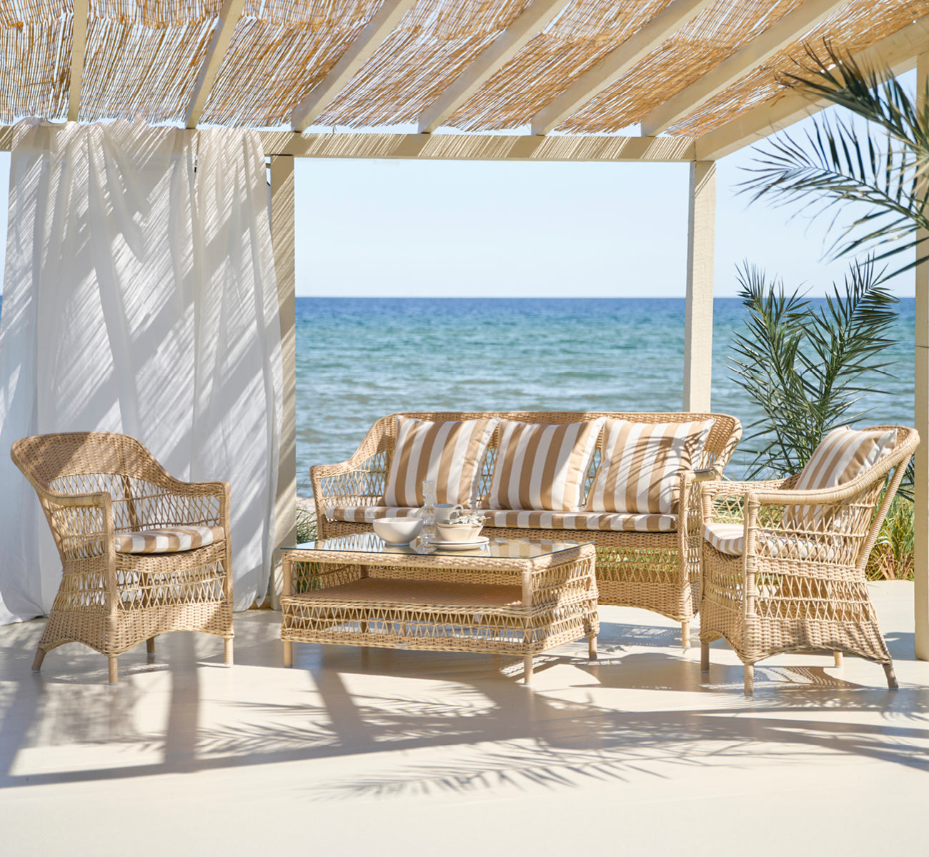 This is a lifestyle image of Hazel Coffee Table in natural ArtFibre Aluminium, on a shaded coastal terrace with reed canopy and sea views.