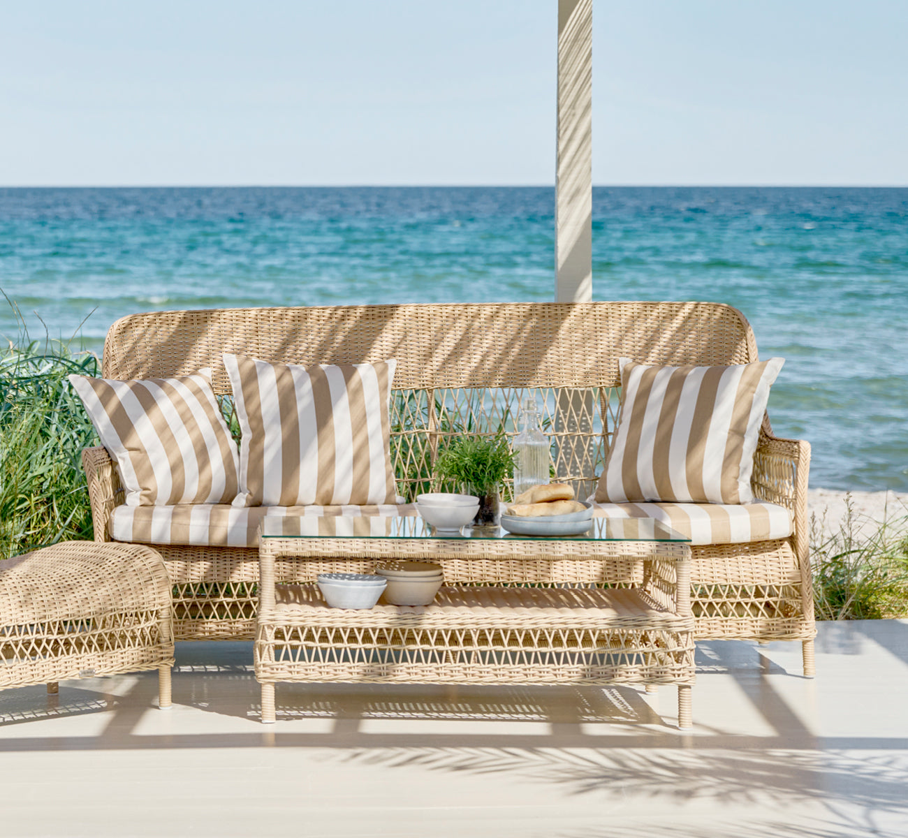 This is a lifestyle image of Hazel Coffee Table in natural ArtFibre Aluminium, on a shaded coastal terrace with reed canopy and sea views.
