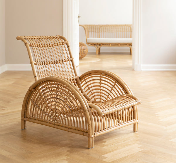 This is a lifestyle image of Paris Lounge Chair in polished natural rattan. Placed on a herringbone parquet floor beside a matching bench.