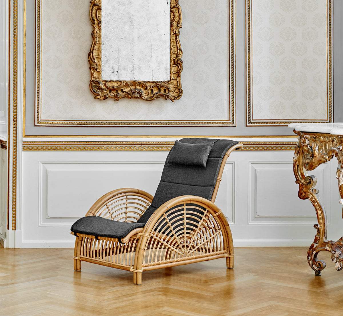 This is a lifestyle image of Paris Lounge Chair in polished natural rattan. Placed on a herringbone parquet floor beside a matching bench.