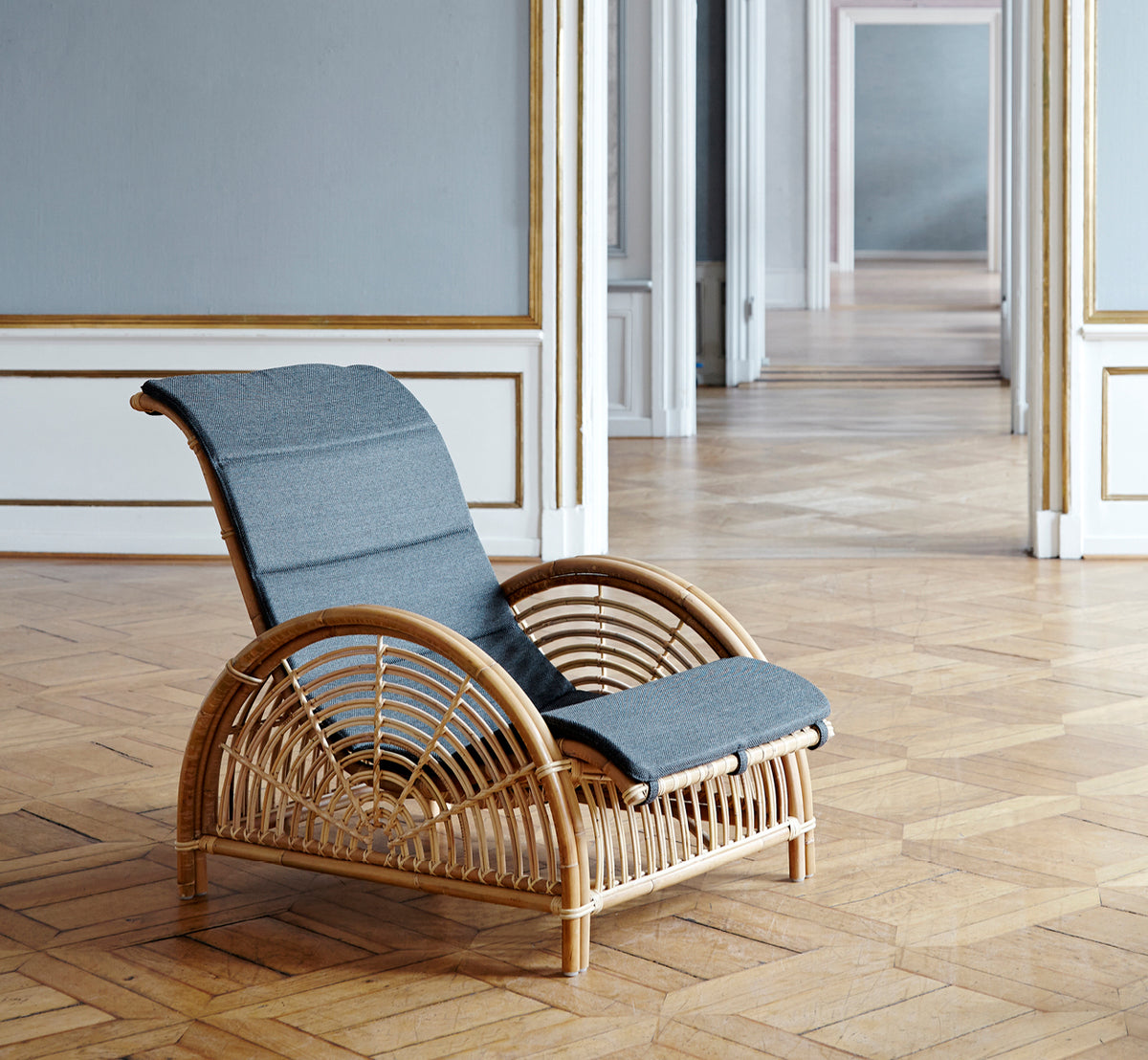 This is a lifestyle image of Paris Lounge Chair in polished natural rattan. Placed on a herringbone parquet floor beside a matching bench.