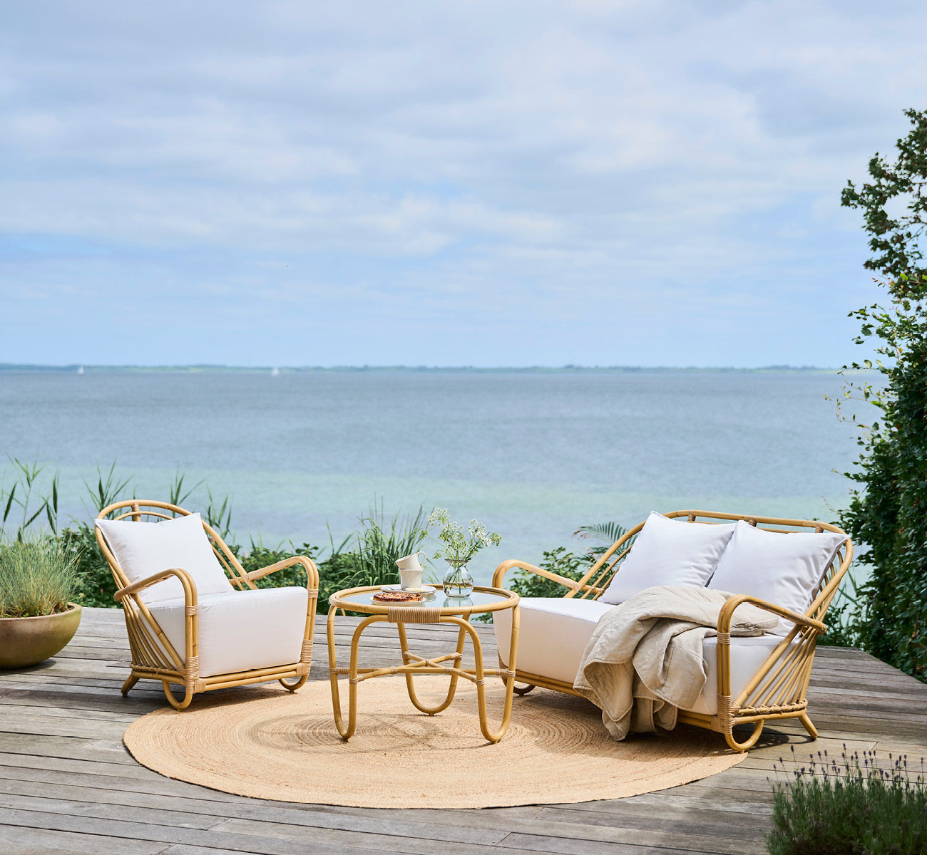 This is a lifestyle image of Charlottenborg Exterior 2 Seater Sofa in natural Aluminium ArtFibre, on an outdoor wooden deck with coastal greenery and.