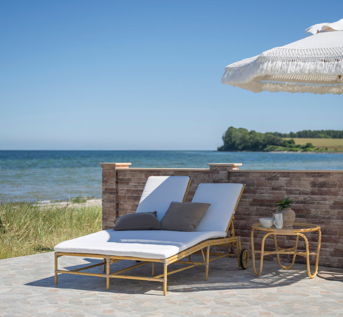This is a lifestyle image of Charlottenborg Exterior Coffee Table in natural aluminium artfibre. Sunbed with cushions under a parasol by the sea.