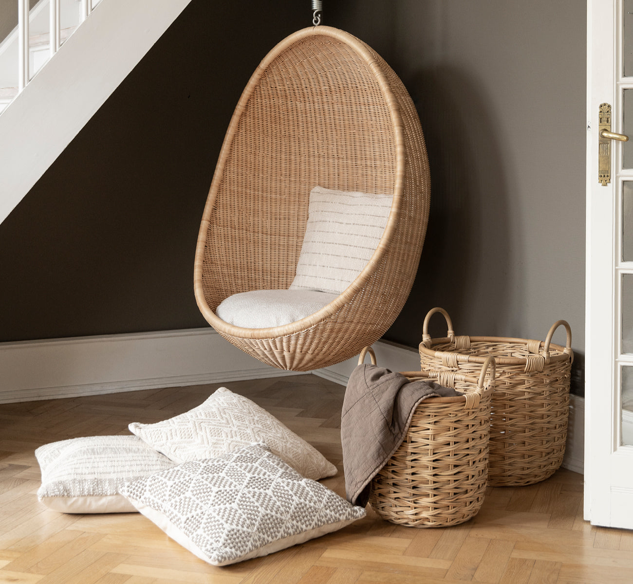 This is a lifestyle image of Sarah Basket set in natural rattan. Placed beside a hanging pod chair and patterned cushions on parquet.