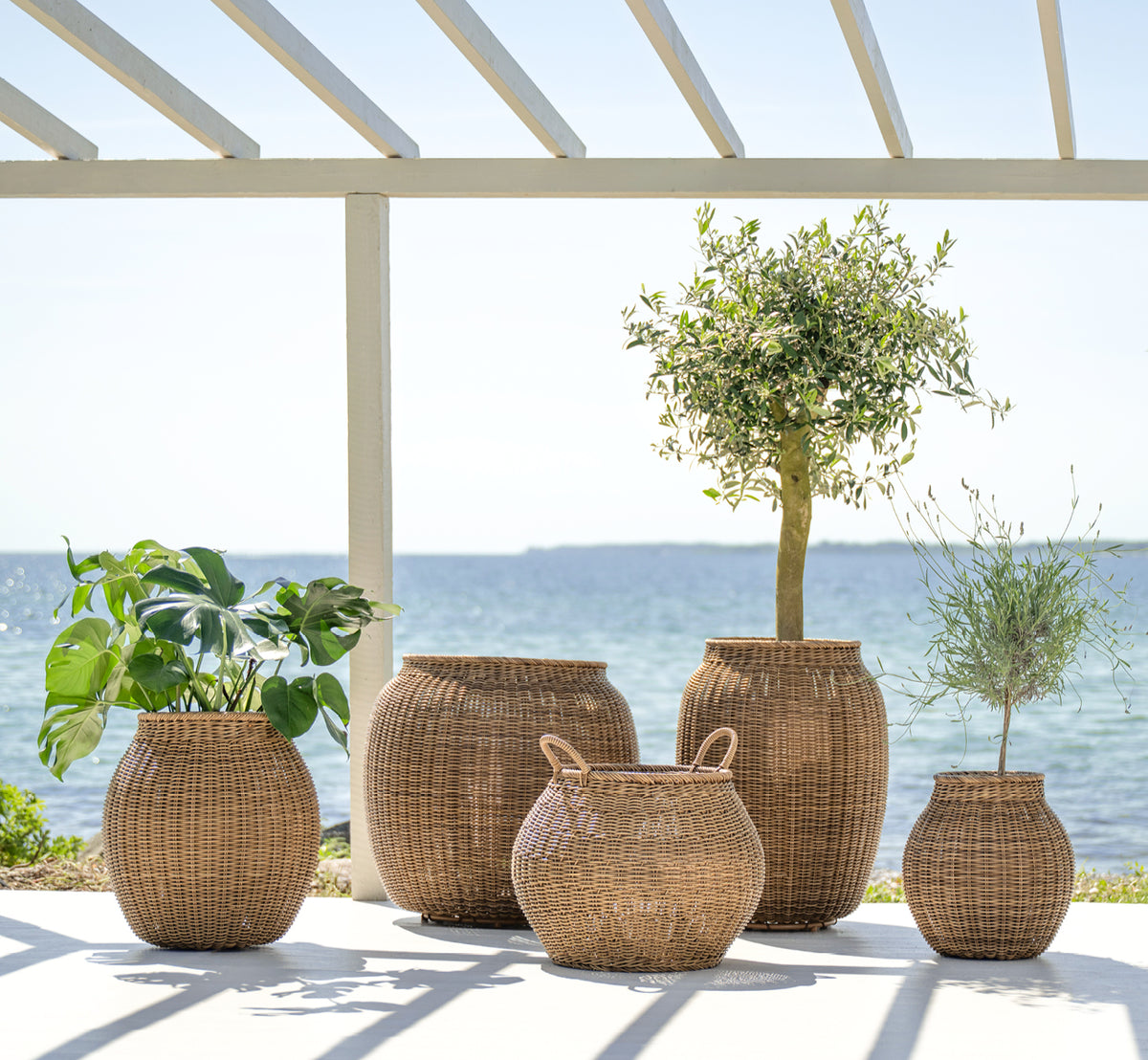 This is a lifestyle image of Ellis Basket in chestnut core weave ArtFibre Aluminium, outdoors on a sun-lit patio with a white wall and tile flooring.