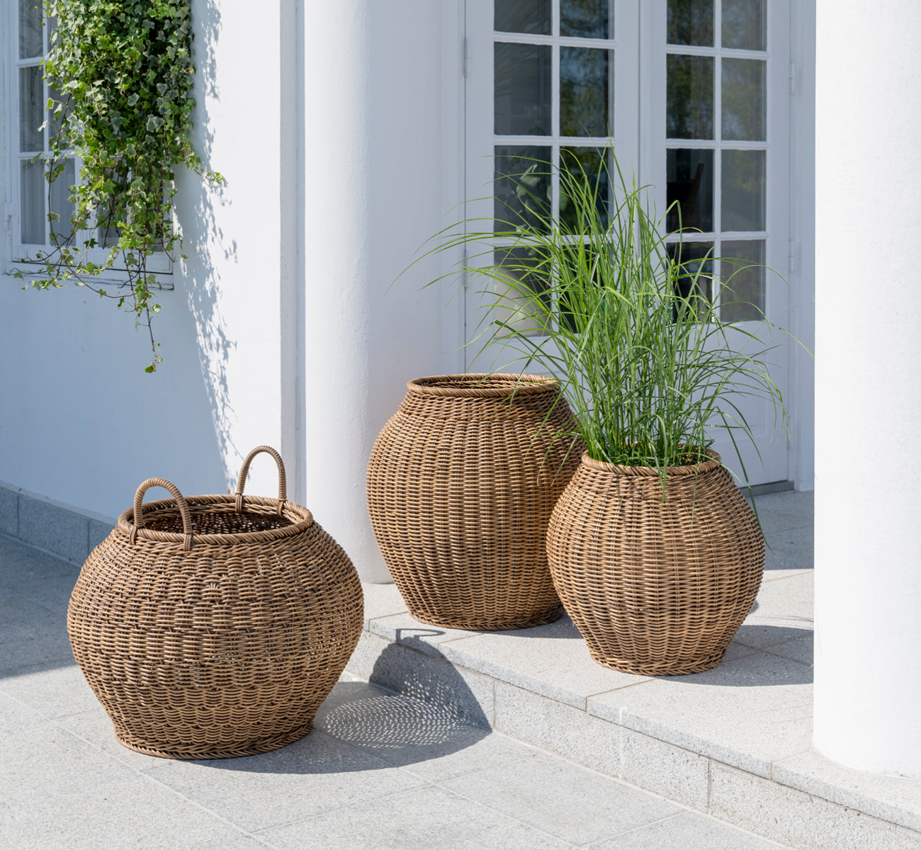 This is a lifestyle image of Ellis Basket in chestnut core weave ArtFibre Aluminium, outdoors on a sun-lit patio with a white wall and tile flooring.