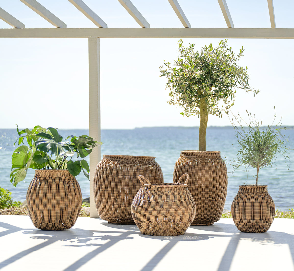 This is a lifestyle image of Ellis Basket in chestnut core weave ArtFibre Aluminium, outdoors on a sun-lit patio with a white wall and tile flooring.