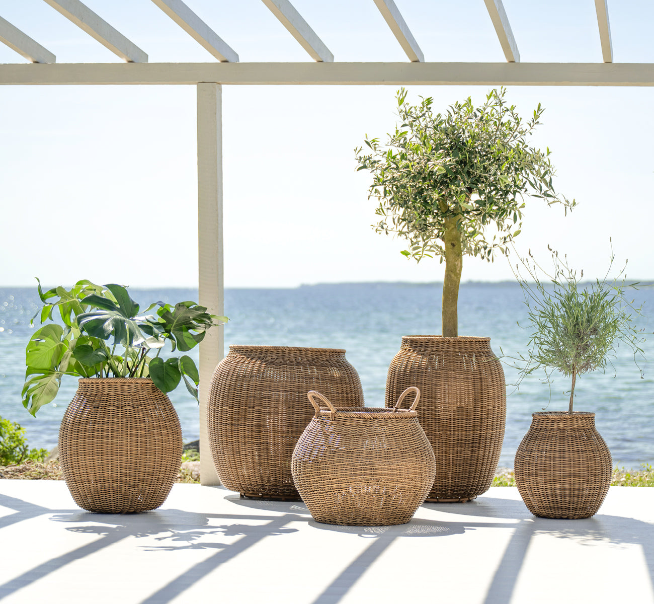 This is a lifestyle image of Ellis Basket in chestnut core weave ArtFibre Aluminium, outdoors on a sun-lit patio with a white wall and tile flooring.