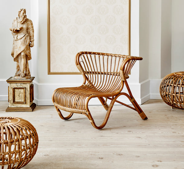 This is a lifestyle image of Ottoman Large in polished antique rattan. Placed in a wallpapered interior with a classical statue backdrop.