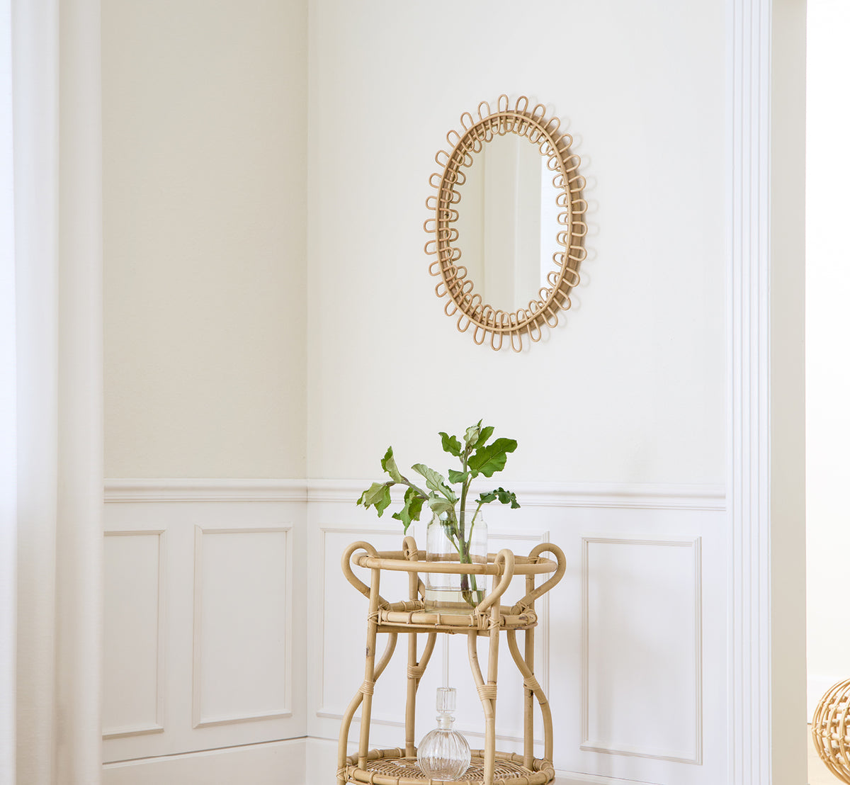 This is a lifestyle image of Luella Mirror in polished natural rattan. Mounted on a white paneled wall above a rattan bar cart with greenery.
