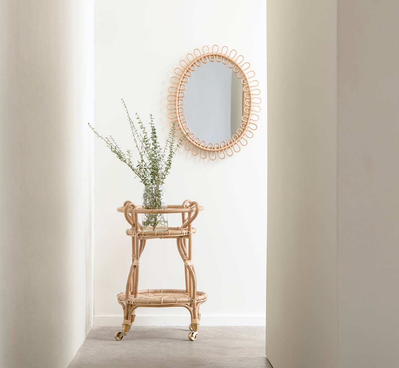This is a lifestyle image of Luella Mirror in polished natural rattan. In a bright hallway alcove above a rattan bar cart with fresh greenery.