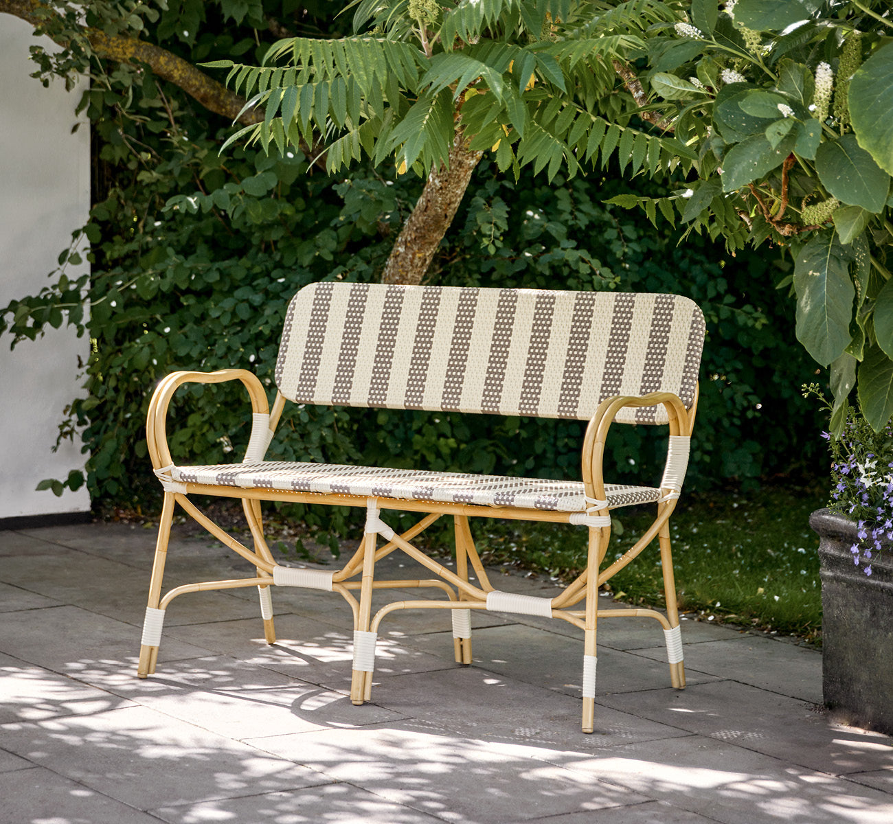 This is a lifestyle image of Bistro Exterior Bench in natural Aluminium ArtFibre, in an outdoor garden café with a bistro table and surrounding trees.