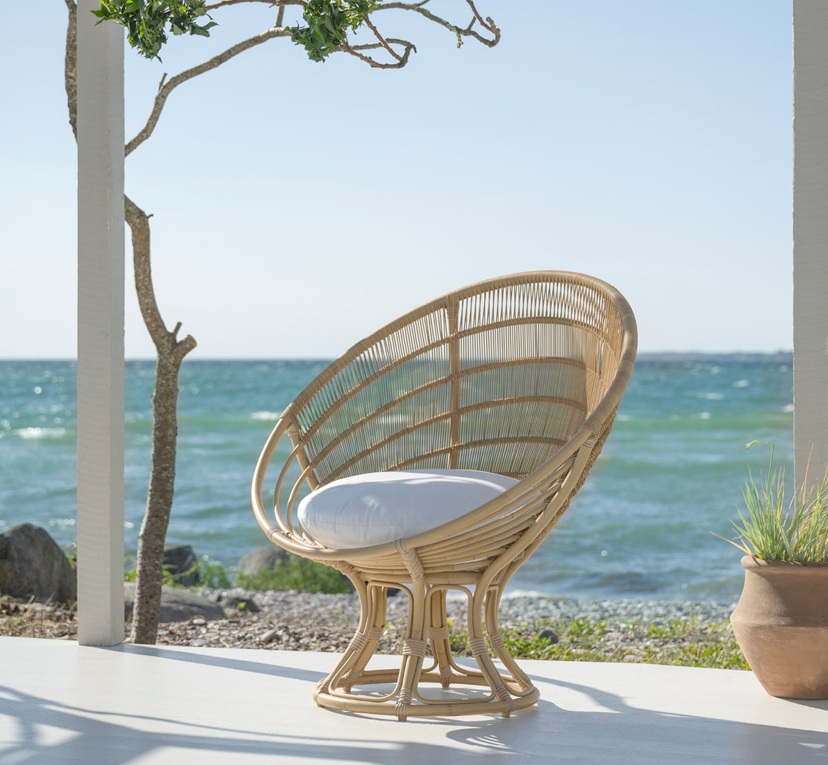 This is a lifestyle image of Luna Exterior Lounge Chair in natural aluminium artfibre. On a white seaside terrace with ocean view and a tree.