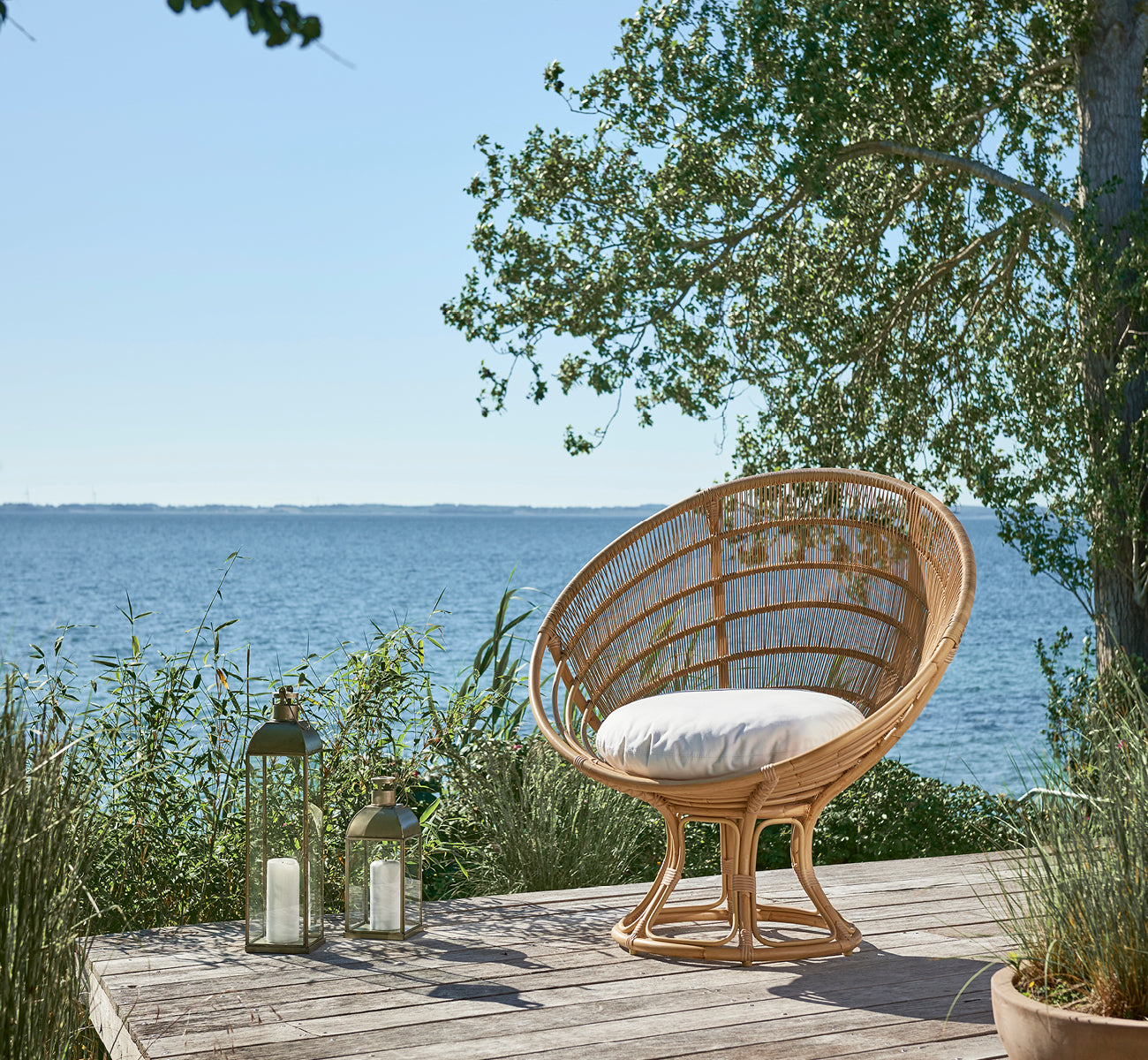 This is a lifestyle image of Luna Exterior Lounge Chair in natural aluminium artfibre. On a wooden deck by the sea surrounded by lush greenery.