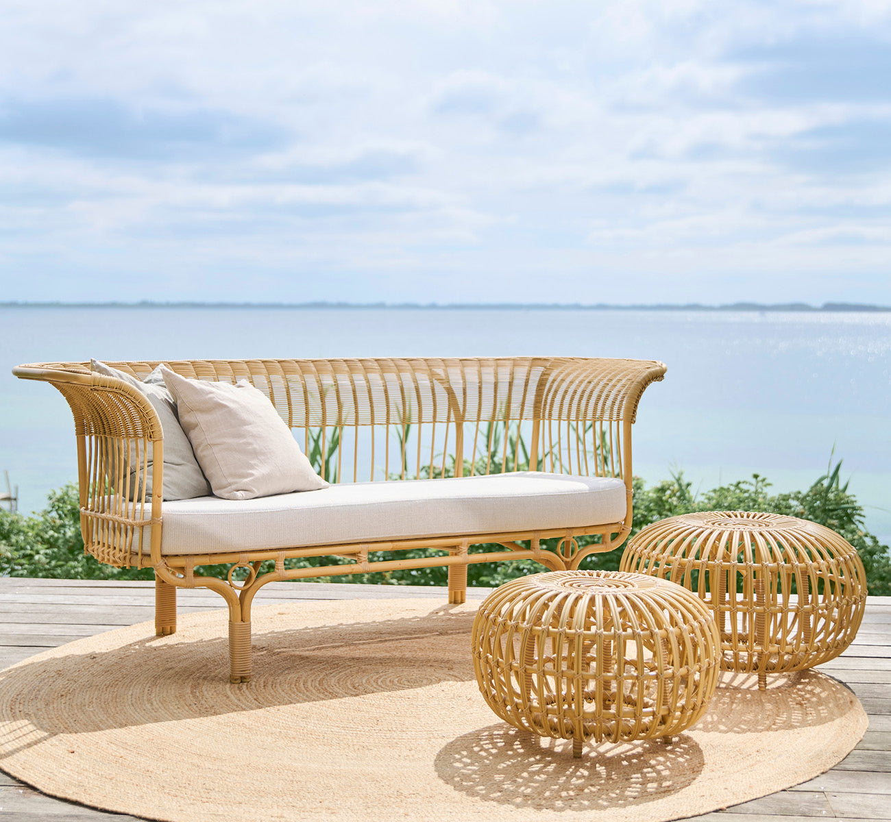 This is a lifestyle image of Belladonna Exterior Sofa in natural Aluminium ArtFibre, on an outdoor wooden deck overlooking the water.