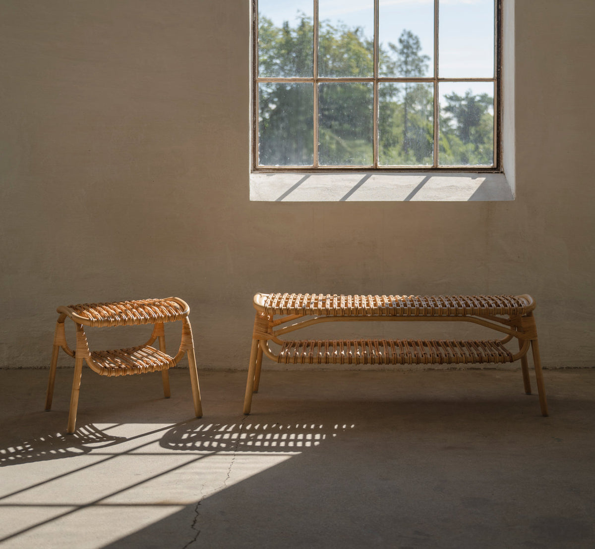 This is a lifestyle image of Machiya Bench in polished natural rattan. With stool by a window in a sunlit room.