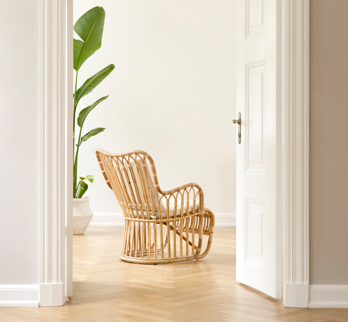 This is a lifestyle image of Tulip Lounge Chair in polished natural rattan. Paired with a matching sofa in a bright panelled room with door.