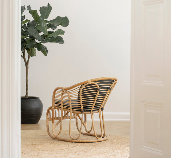 This is a lifestyle image of Swing Lounge Chair in polished natural rattan. Standing alone beside a leafy plant on a herringbone parquet floor.