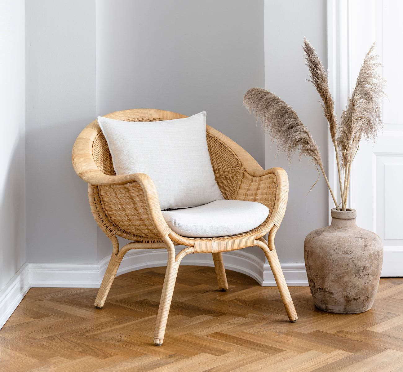 This is a lifestyle image of Madame Lounge Chair in natural core weave rattan frame. Cozy corner with herringbone floor and pampas grass vase.