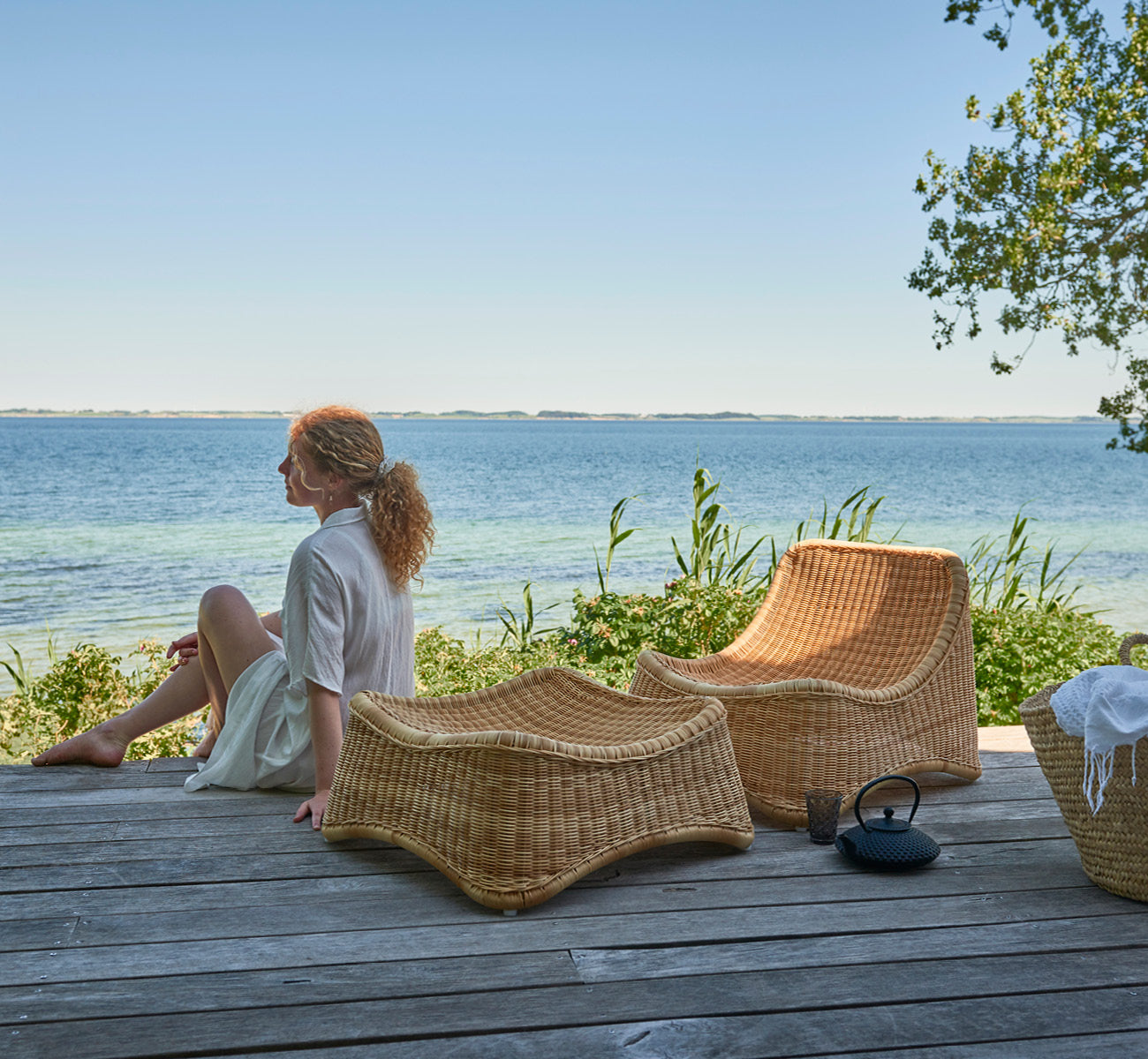 This is a lifestyle image of Chill Lounge Chair in natural core weave ArtFibre Aluminium, on a waterside wooden deck with coastal greenery and sea.