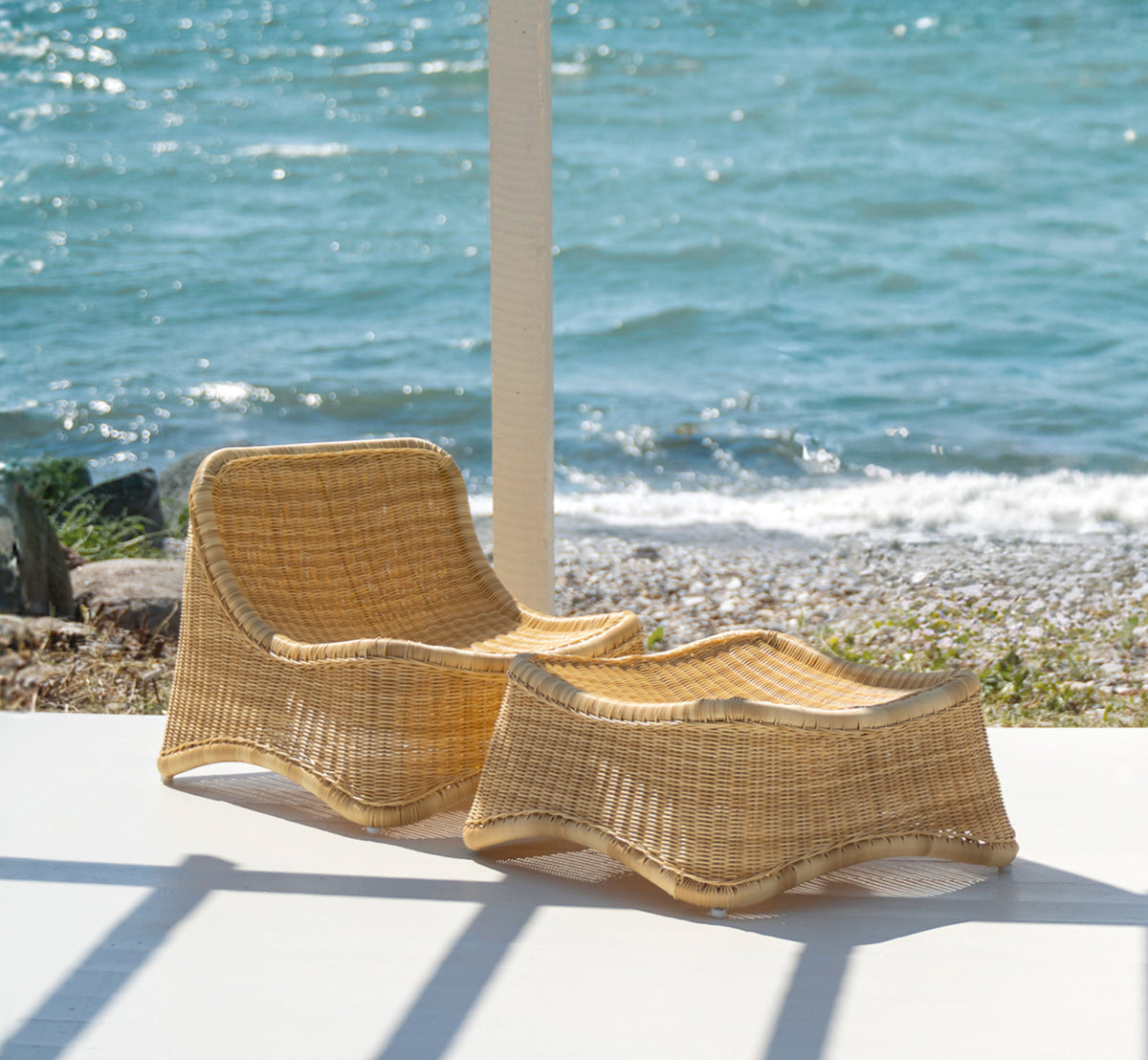 This is a lifestyle image of Chill Lounge Chair in natural core weave ArtFibre Aluminium, on a waterside wooden deck with coastal greenery and sea.