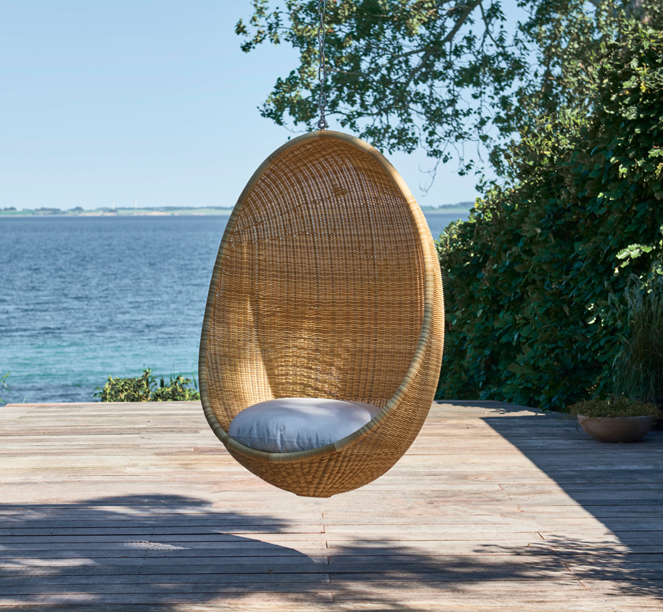 This is a lifestyle image of Hanging Egg Exterior Chair in natural core weave ArtFibre Aluminium, hanging from a white pergola with sandy beach and.
