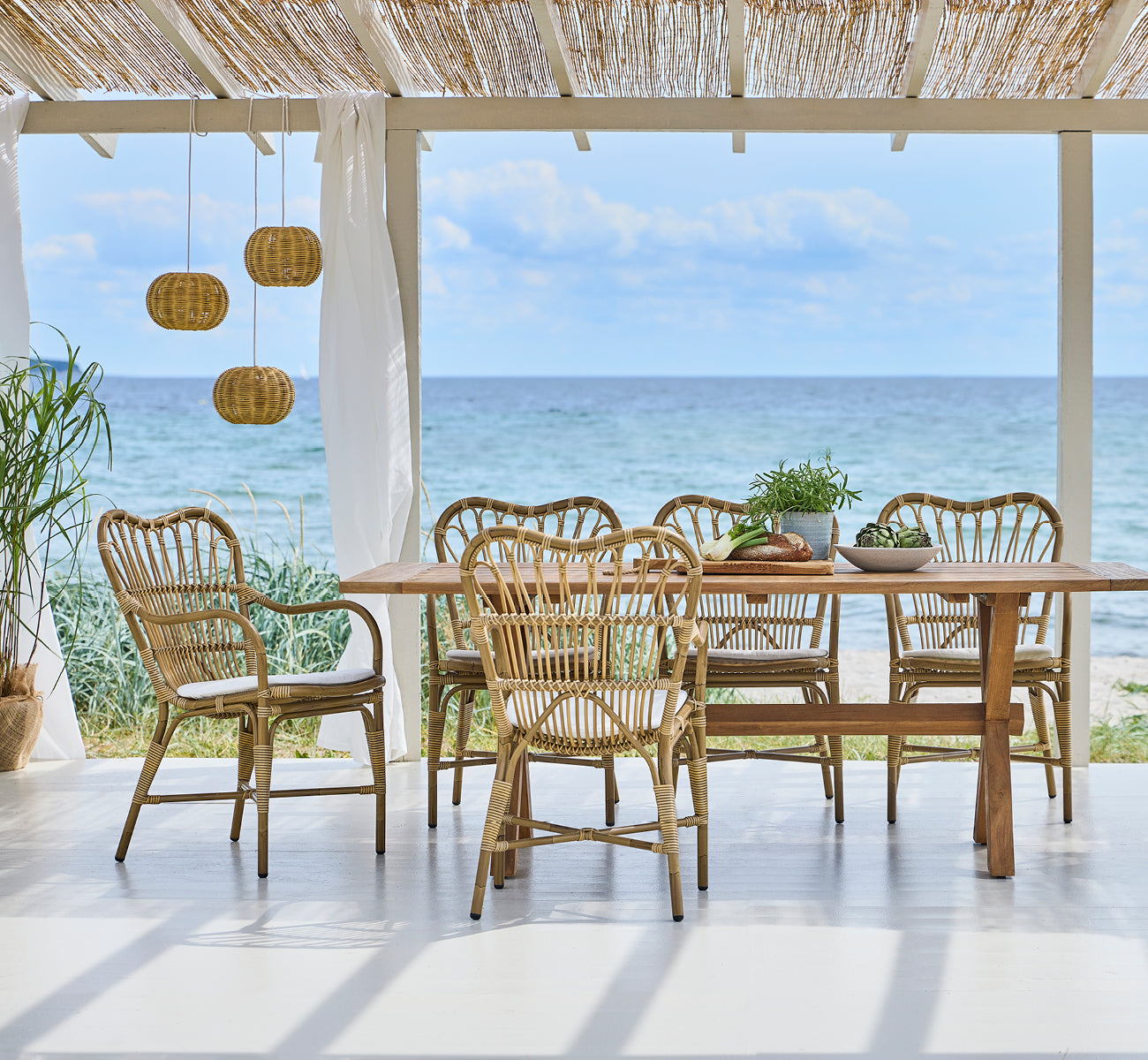 This is a lifestyle image of Margret Exterior Dining Armchair in antique aluminium artfibre. Beach pavilion with pendant lamps and ocean view.
