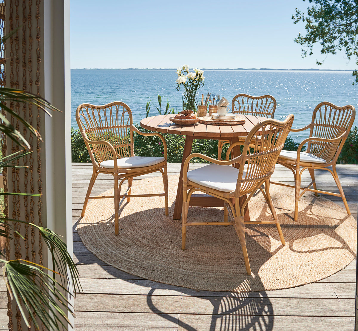This is a lifestyle image of Margret Exterior Dining Armchair in natural aluminium artfibre. Seaside deck with round teak table and ocean view.