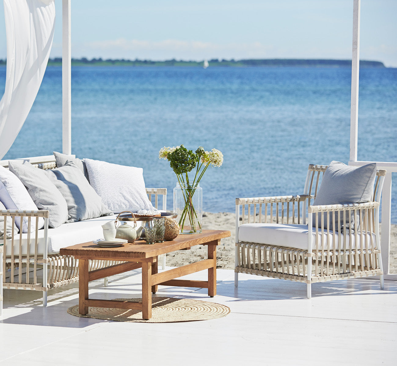 This is a lifestyle image of Caroline Exterior Lounge Chair in dove White Aluminium ArtFibre, on a waterside wooden deck with coastal greenery and.