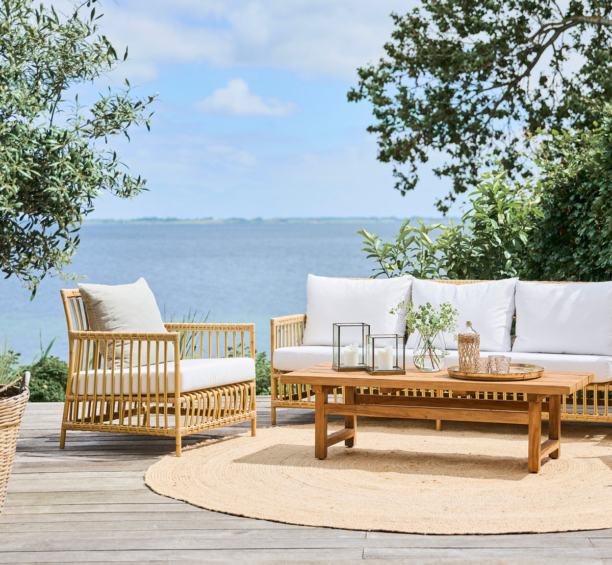 This is a lifestyle image of Caroline Exterior Lounge Chair in natural Aluminium ArtFibre, on a waterside wooden deck with coastal greenery and sea.