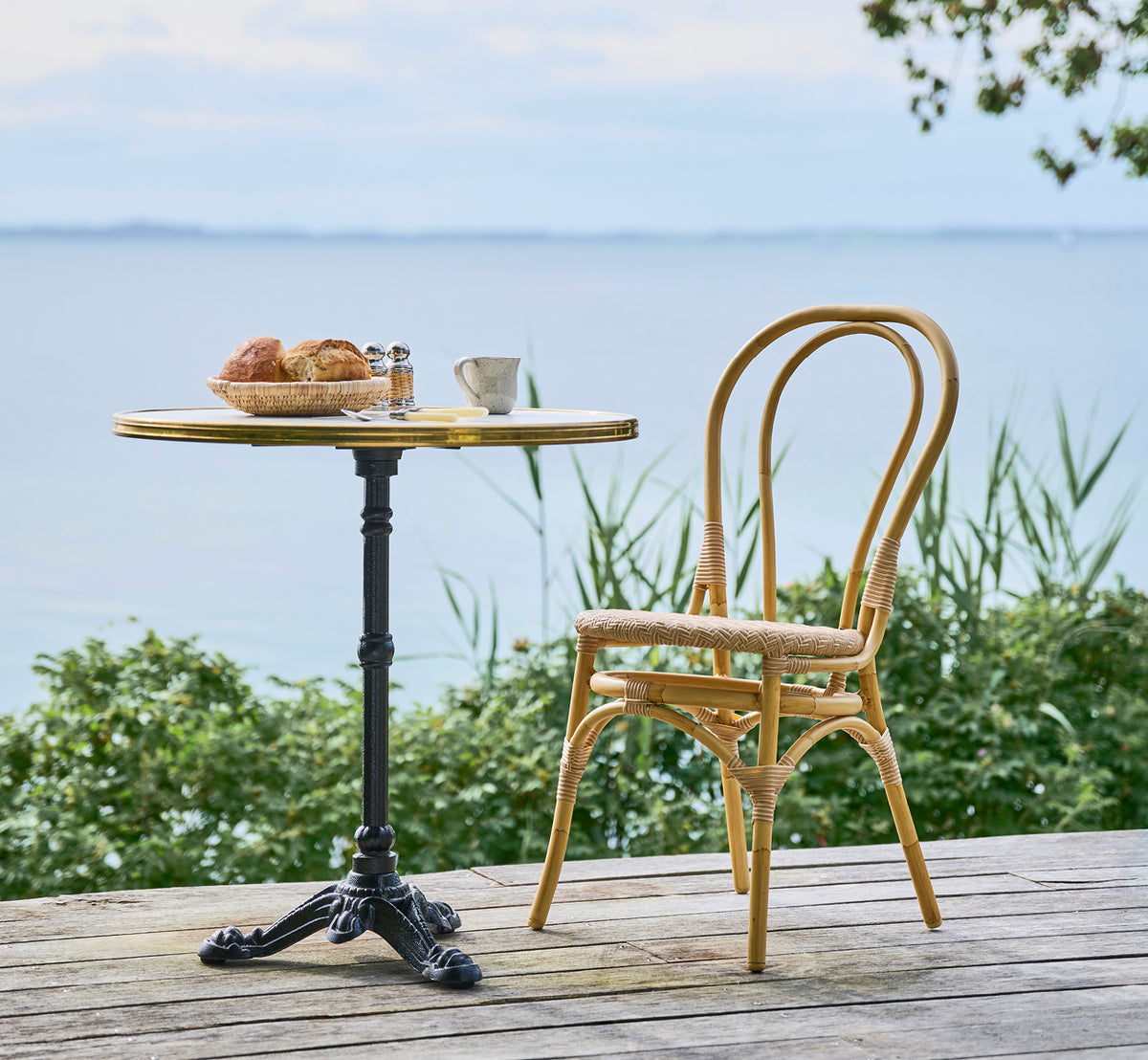 This is a lifestyle image of Lulu Exterior Dining Chair in natural aluminium artfibre. At a waterfront bistro table with pastries and calm sea.