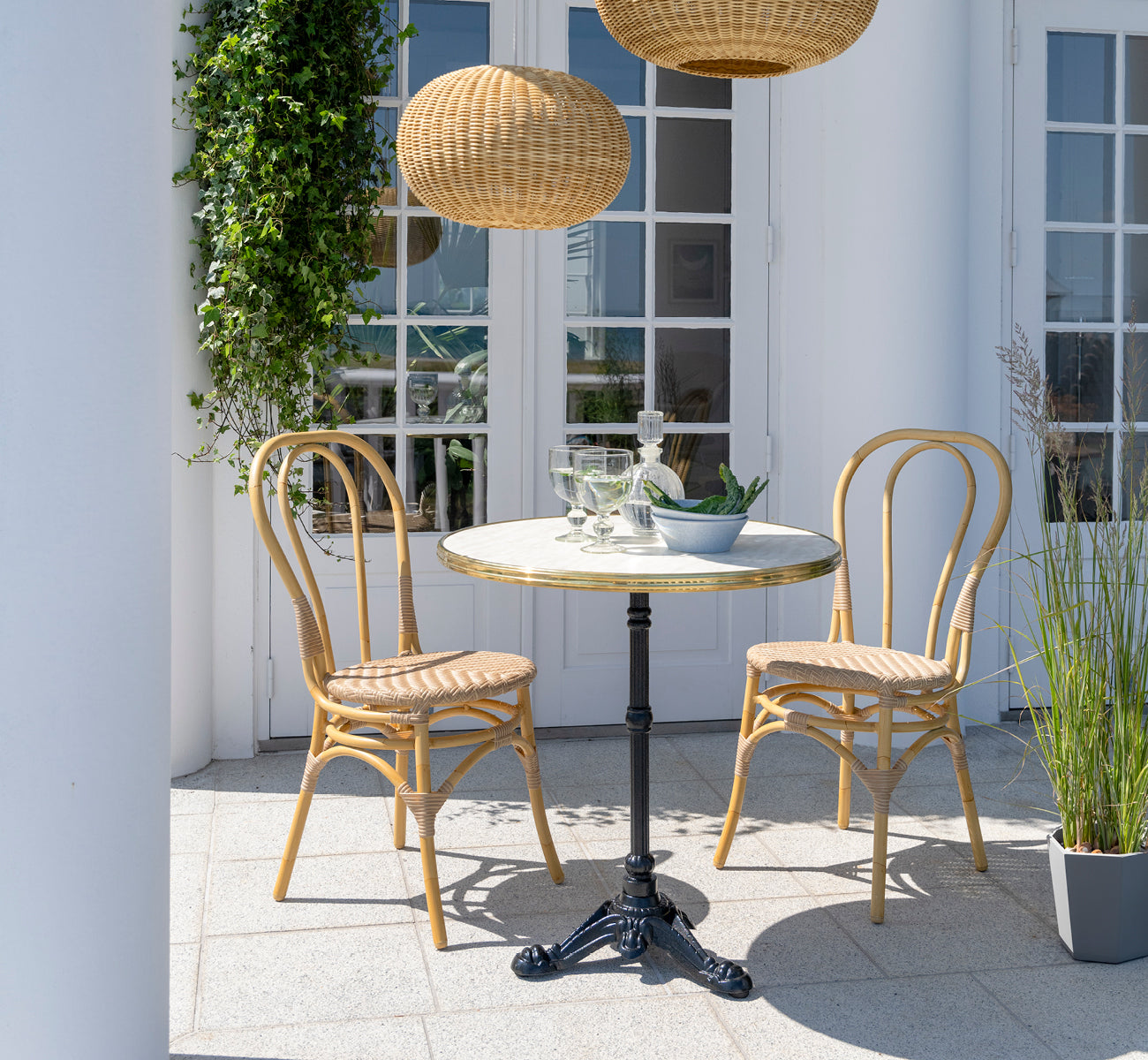 This is a lifestyle image of Lulu Exterior Dining Chair in natural aluminium artfibre. On a white terrace with a café table and climbing plants.