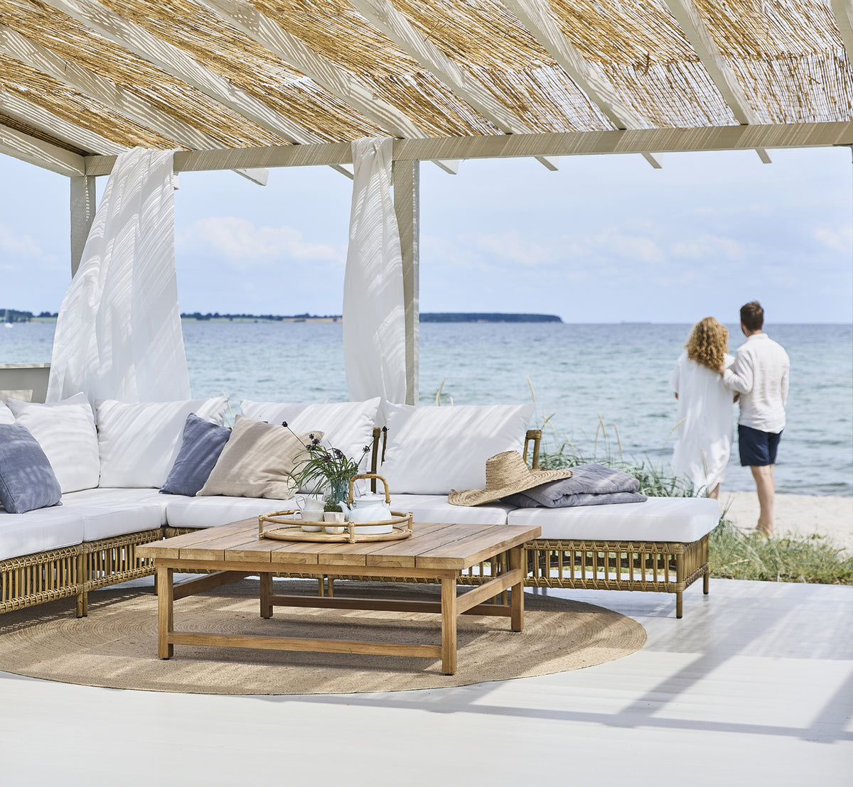 This is a lifestyle image of Maggie Sofa Pouf Module w. Glass in antique aluminium artfibre. Beach pavilion with couple and ocean view.