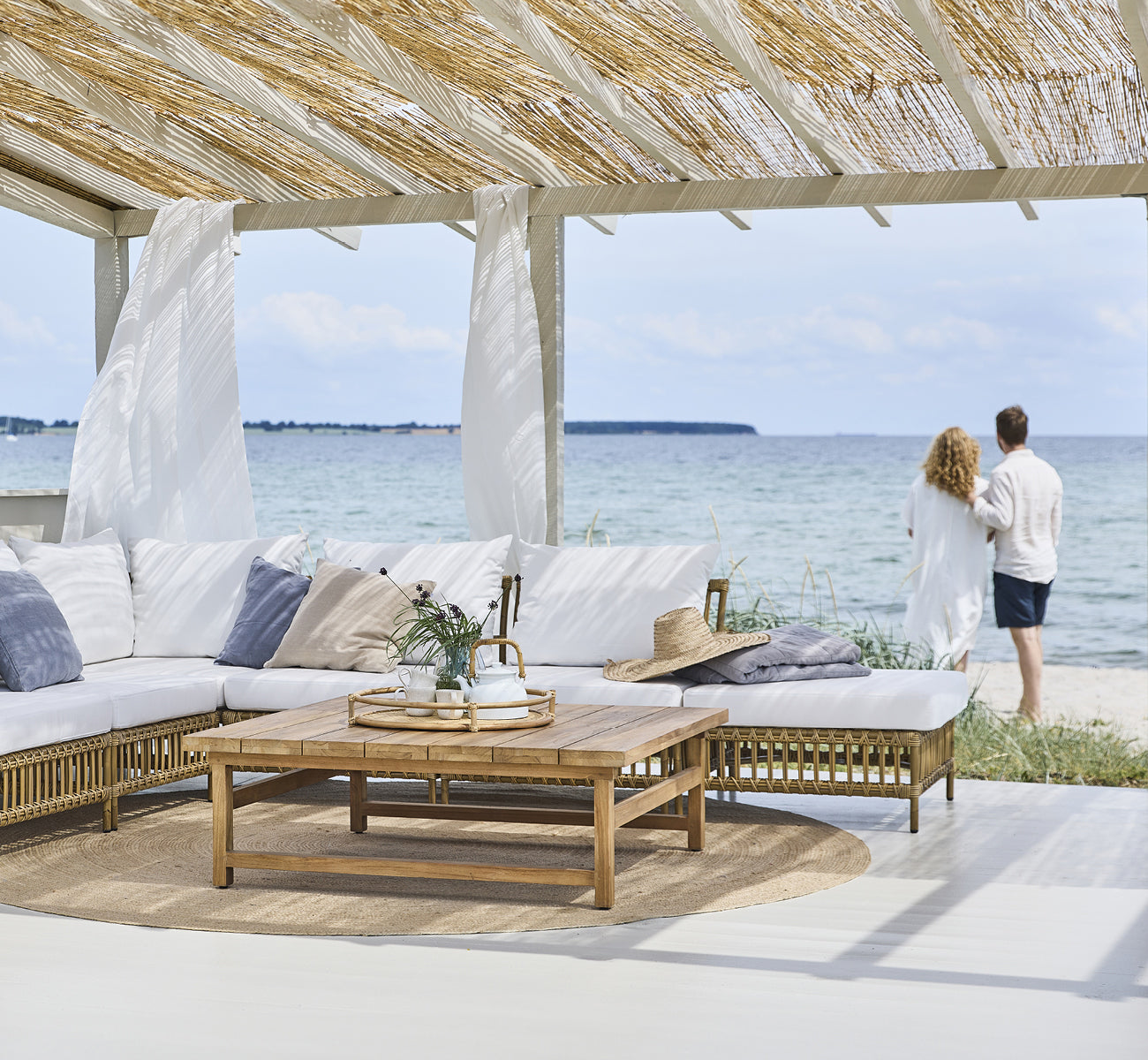This is a lifestyle image of Maggie Sofa Pouf Module w. Glass in antique aluminium artfibre. Beach pavilion with couple and ocean view.