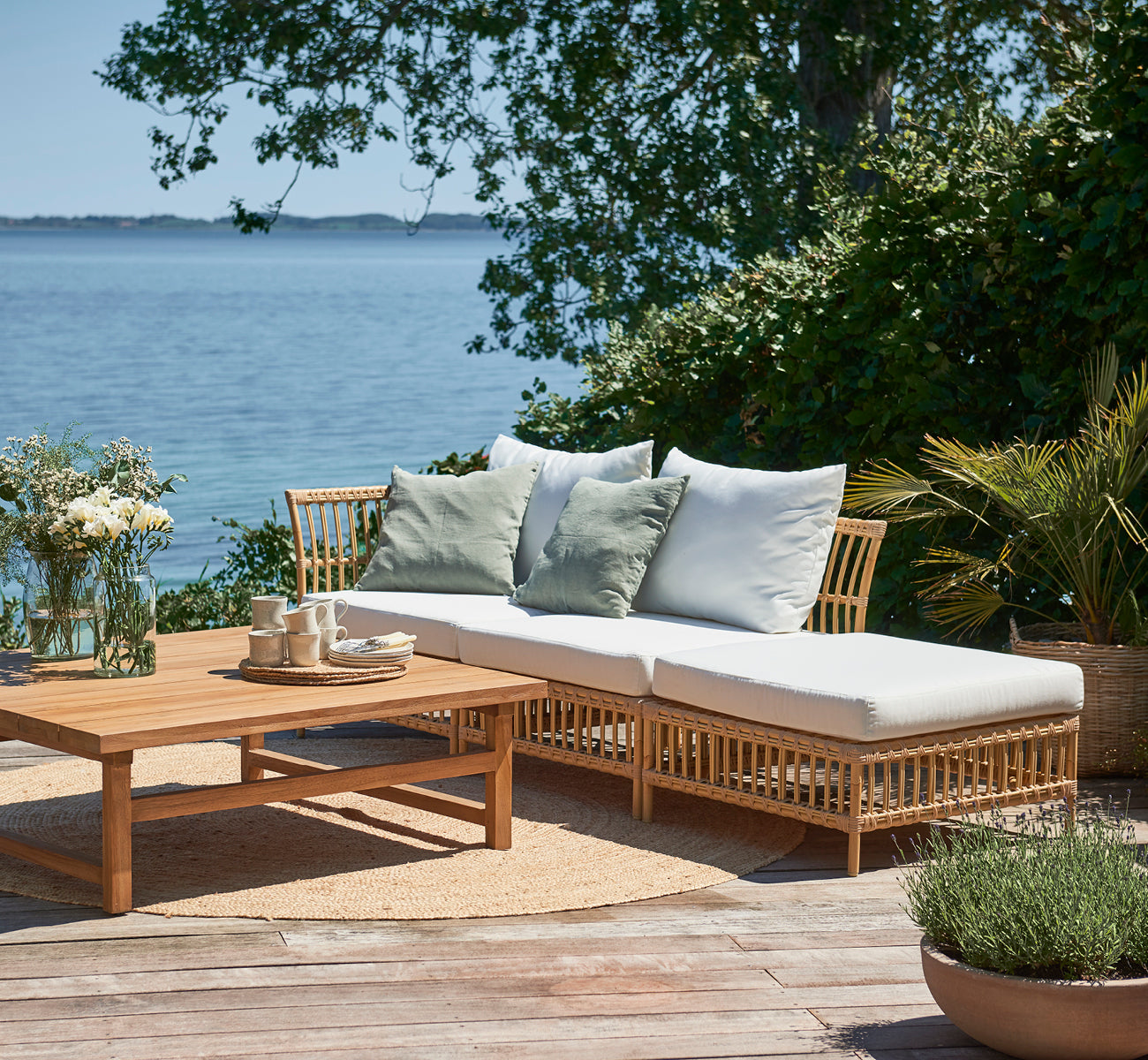 This is a lifestyle image of Maggie Sofa Pouf Module w. Glass in natural aluminium artfibre. Seaside deck with coffee table and green cushions.