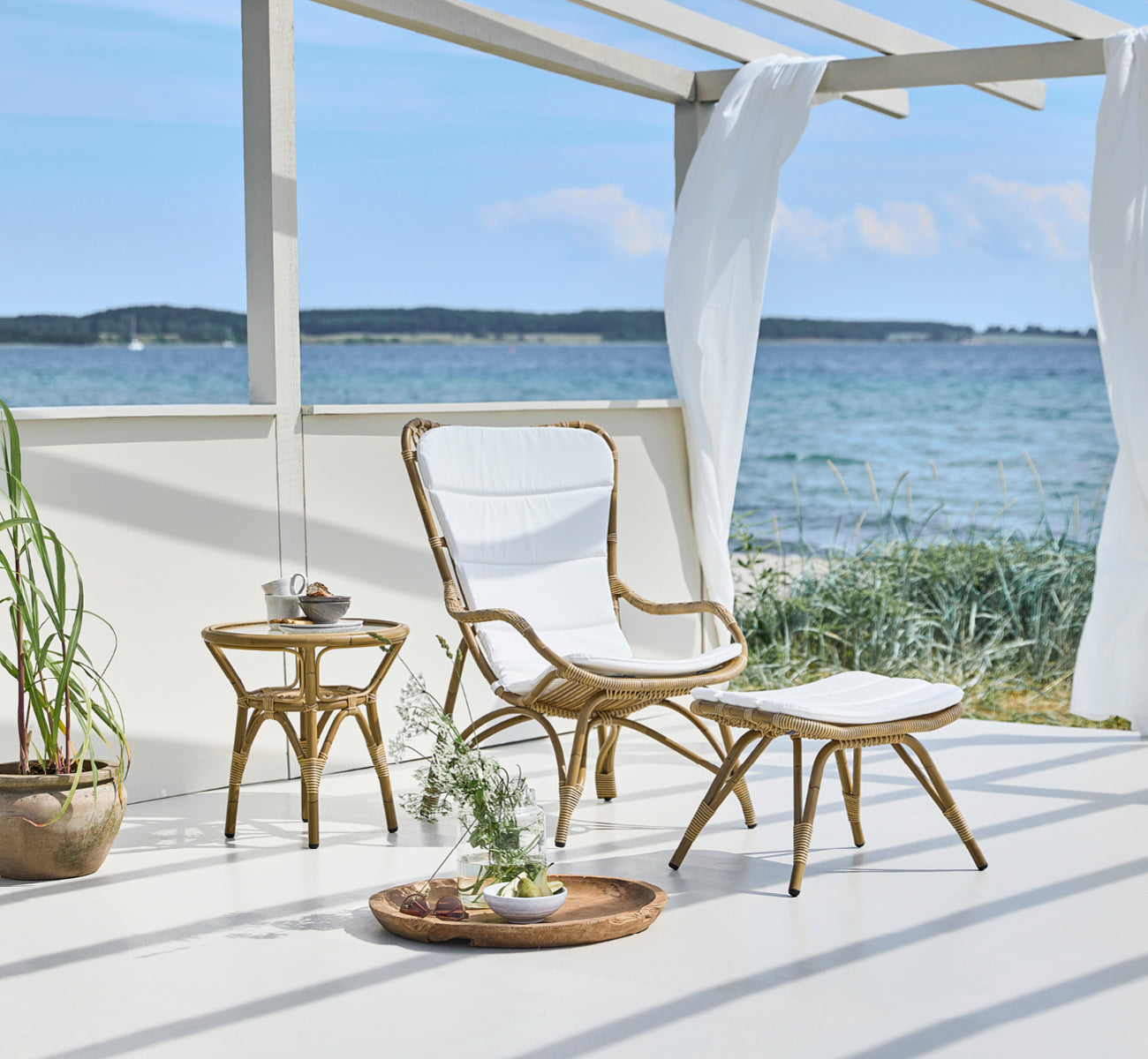This is a lifestyle image of Monet Exterior Footstool in antique aluminium artfibre. On a seaside pavilion with cushioned chair.