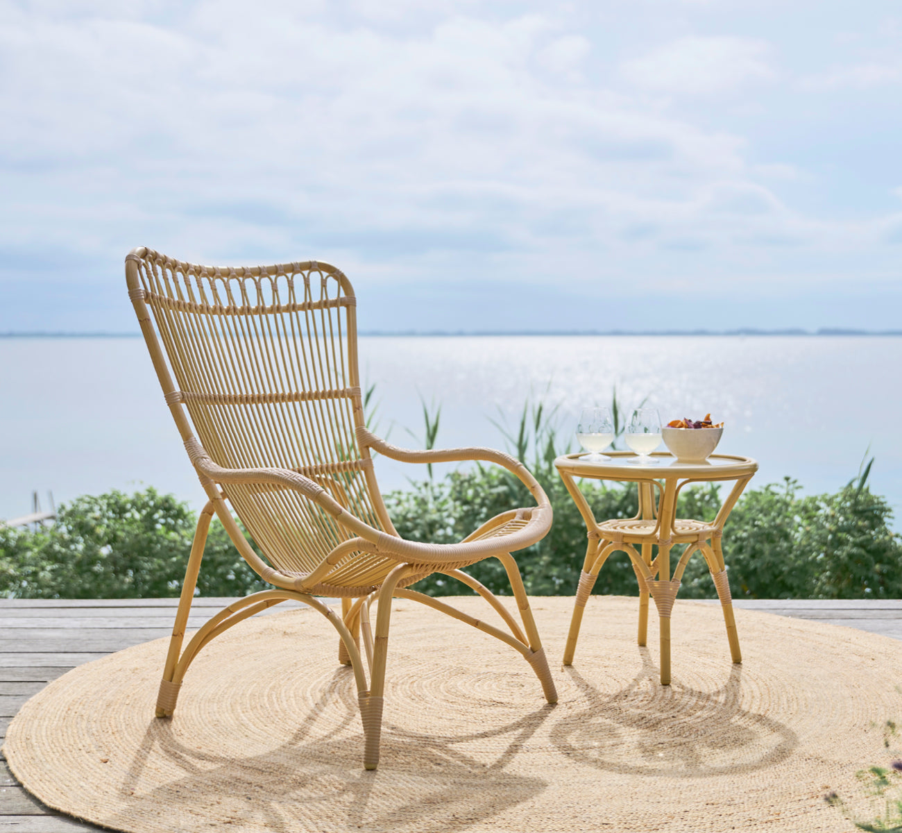 This is a lifestyle image of Tony Exterior Side Table in natural aluminium artfibre. Placed beside a high-back rattan chair on a sunlit wooden deck.