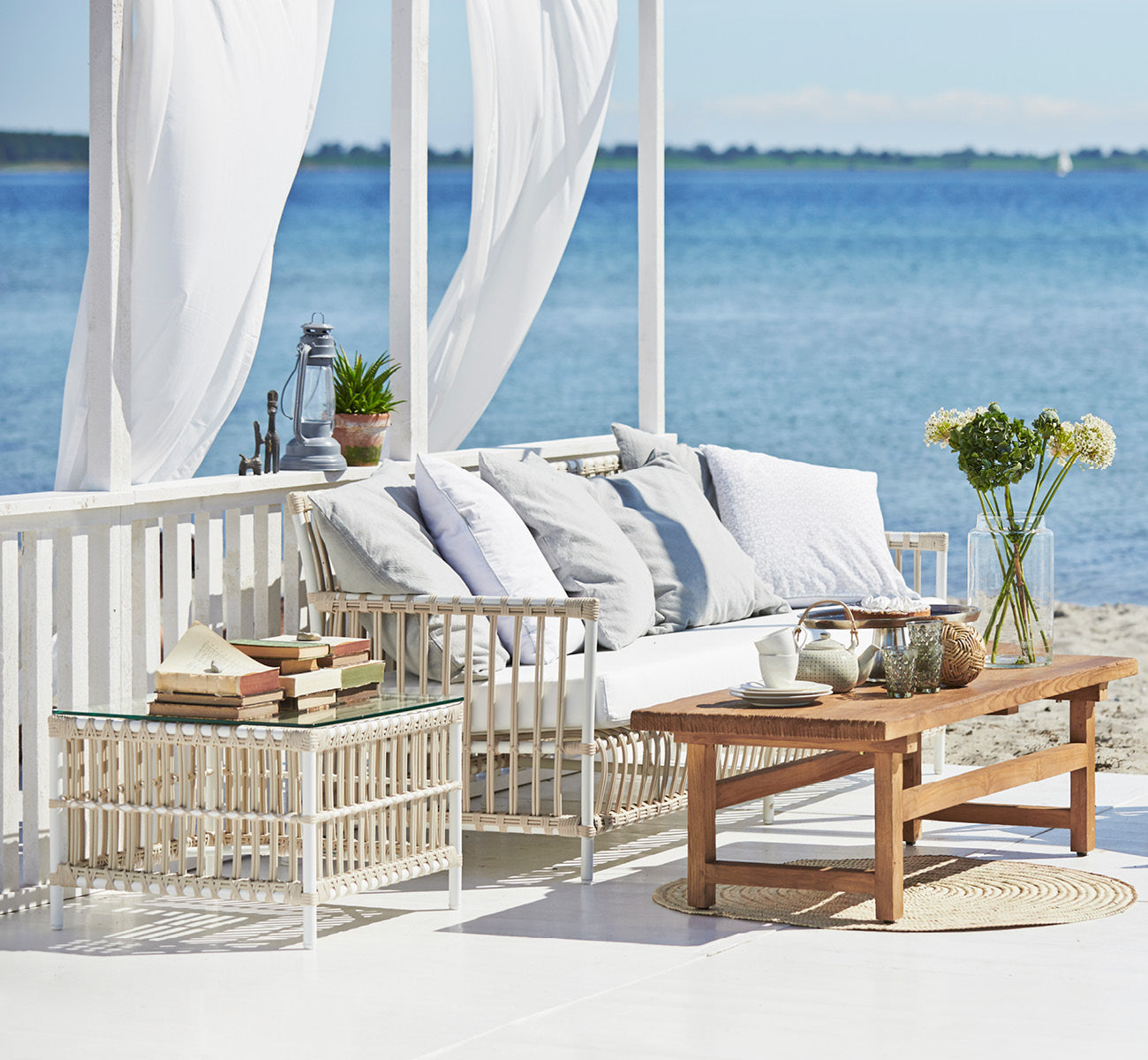 This is a lifestyle image of Caroline Exterior Table in dove White Aluminium ArtFibre, on a waterside wooden deck with coastal greenery and sea views.