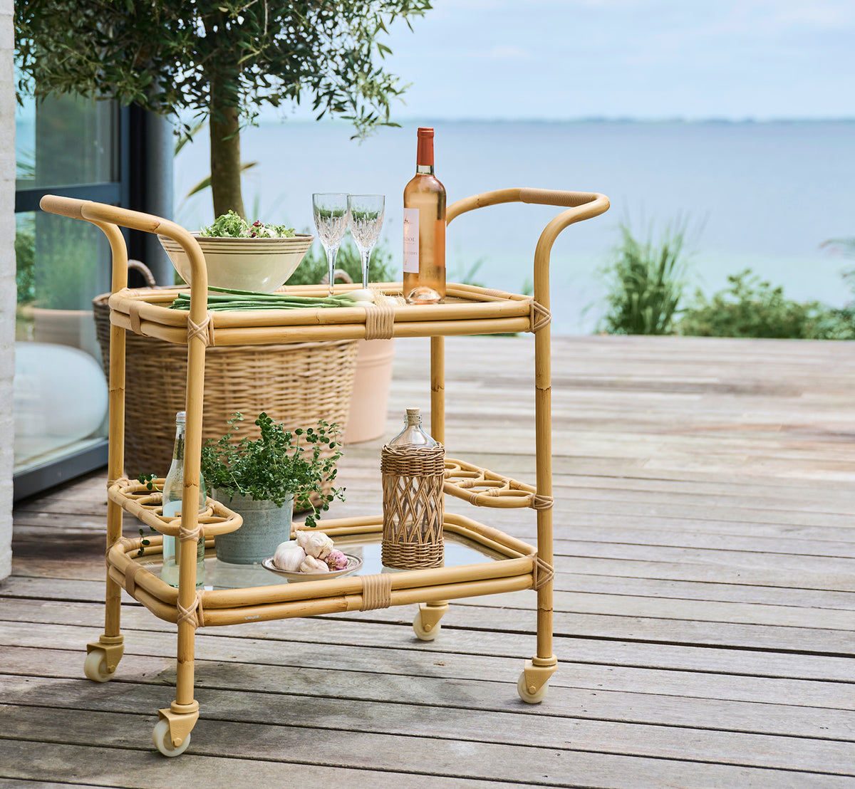 This is a lifestyle image of Carlo Exterior Trolley in natural Aluminium ArtFibre, on a waterside wooden deck with coastal greenery and sea views.