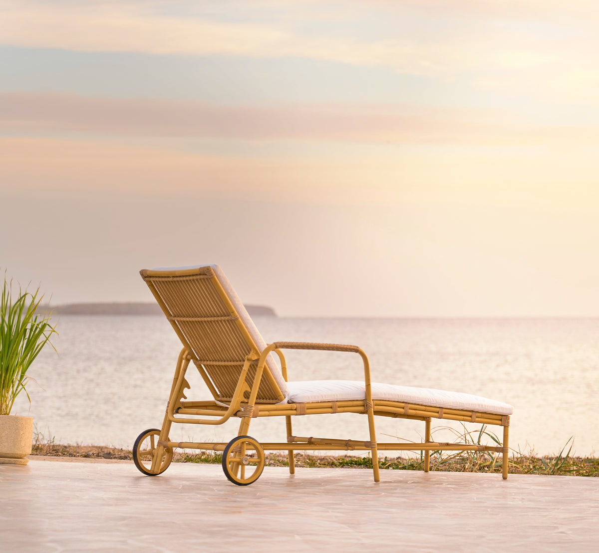 This is a lifestyle image of Josephine sunbed in natural aluminium artfibre, on a coastal stone terrace at sunset with sea views and beach grasses.