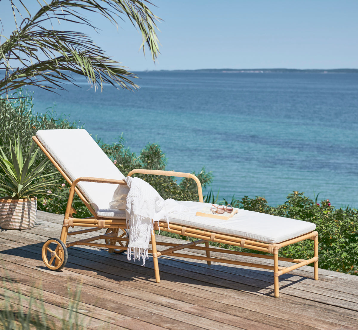 This is a lifestyle image of Josephine sunbed in natural aluminium artfibre, on a coastal stone terrace at sunset with sea views and beach grasses.