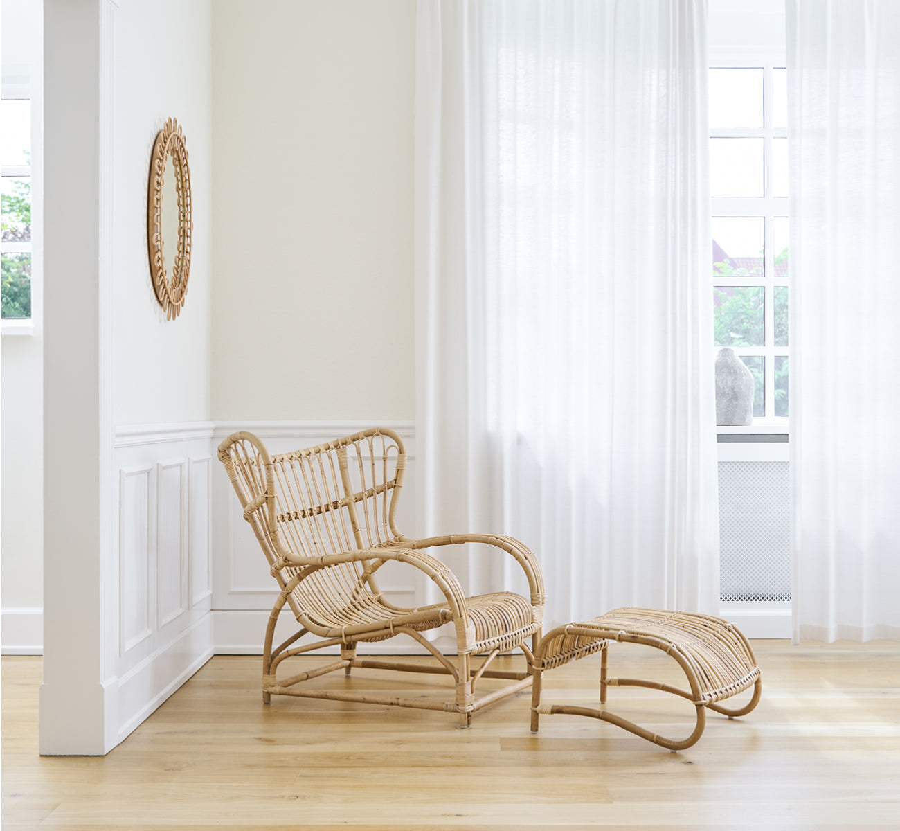 This is a lifestyle image of Teddy Lounge Chair in polished natural rattan. Placed on a herringbone floor by a curtained window with small mirror.