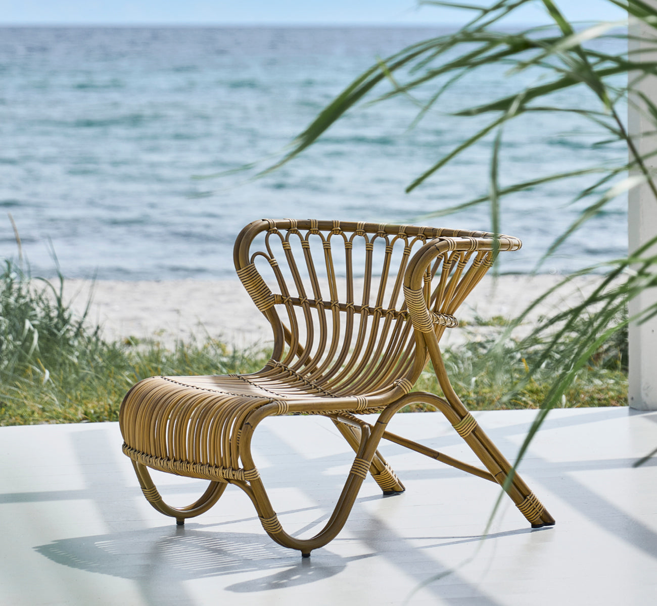 This is a lifestyle image of Fox Exterior Lounge Chair in antique Aluminium ArtFibre, on a waterside wooden deck with coastal reeds and sea views.