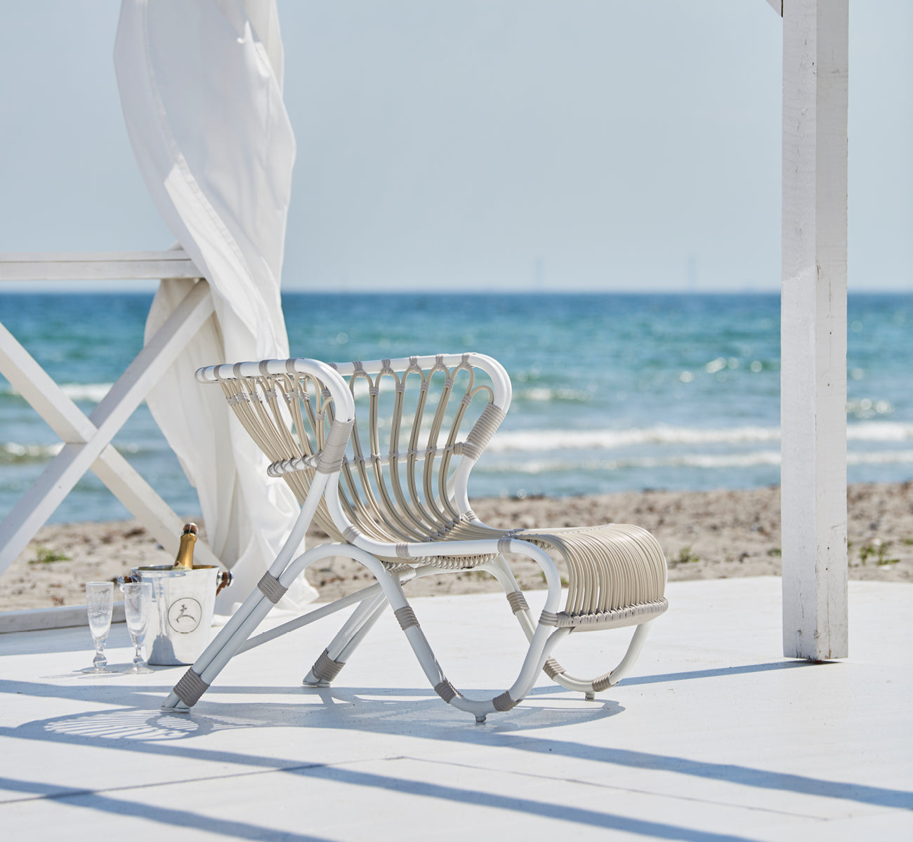 This is a lifestyle image of Fox Exterior Lounge Chair in dove white Aluminium ArtFibre, on a waterside wooden deck with coastal reeds and sea views.
