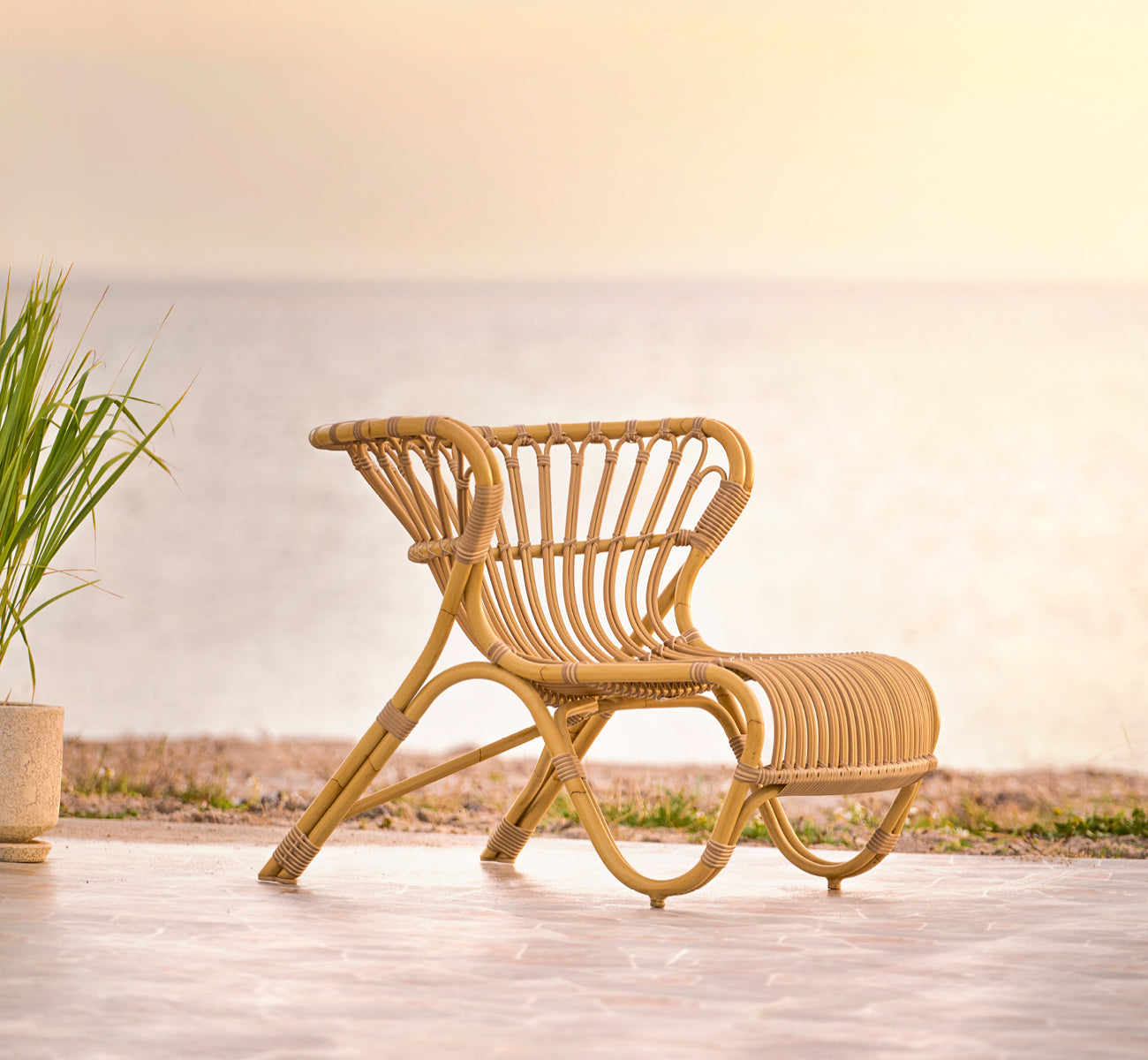 This is a lifestyle image of Fox Exterior Lounge Chair in natural Aluminium ArtFibre, on a waterside wooden deck with coastal reeds and sea views.