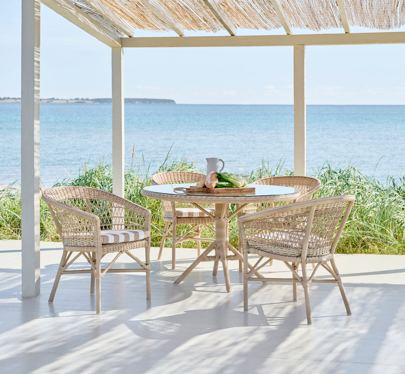 This is a lifestyle image of Emma Exterior Dining Chair in natural ArtFibre Aluminium, on a bright coastal terrace with a reed roof shade and sea.