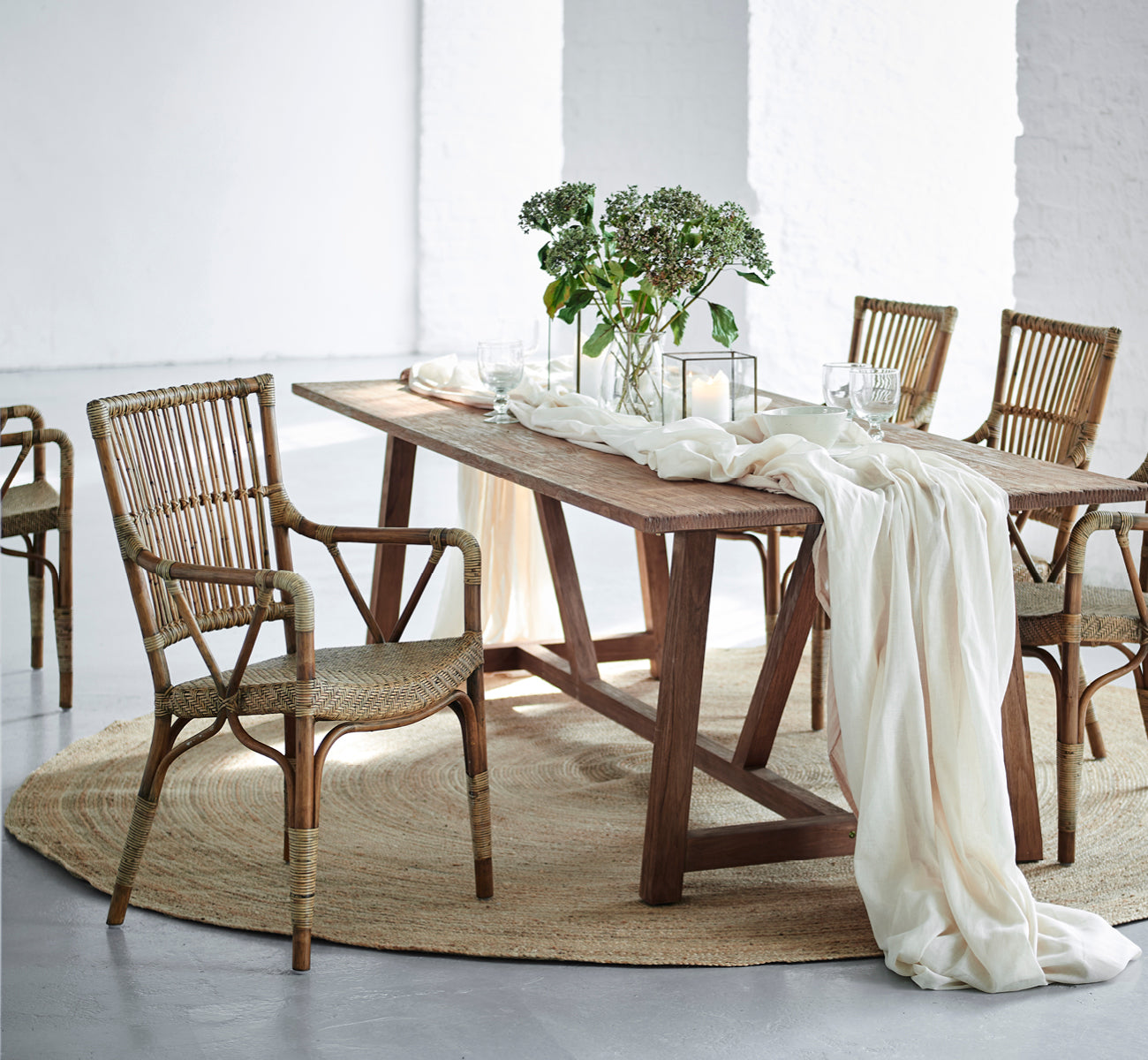 This is a lifestyle image of Jute Carpet Ø280 in grey jute. Beneath a dining table with rattan chairs, linen draping, and greenery.