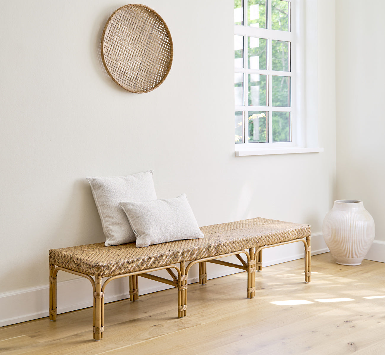 This is a lifestyle image of Luis Bench in antique rattan. Styled with linen cushions in a bright white room with wall décor and a white vase.